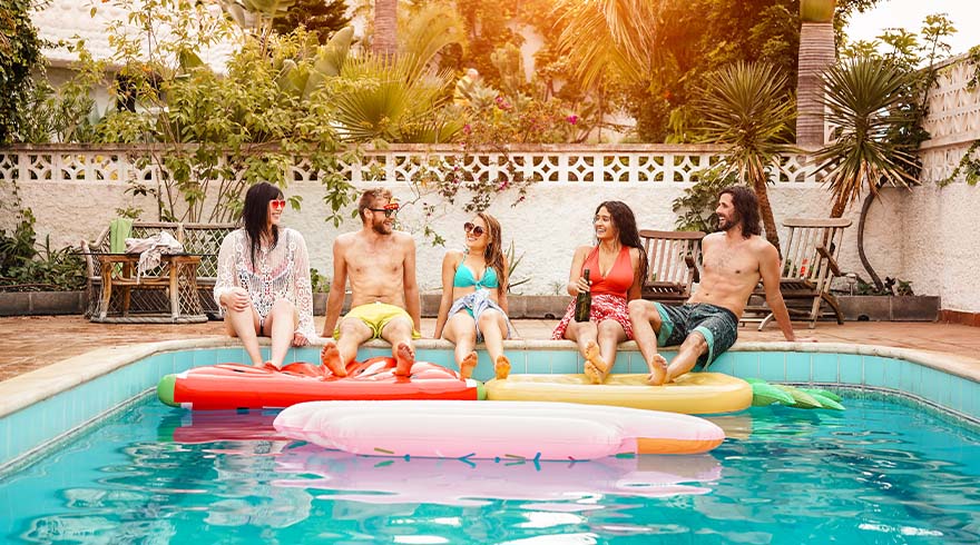 Vijf mensen zitten aan de rand van een zwembad met opblaasbare figuren. De achtergrond toont palmen en een witte muur.