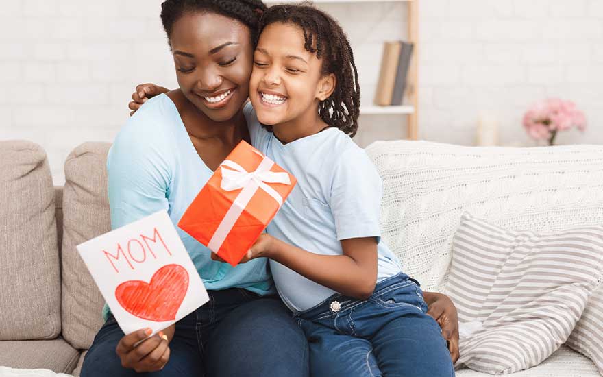 Mother and daughter cuddle on the sofa, smiling widely while exchanging a red gift box and a handmade card marked “MOM”.