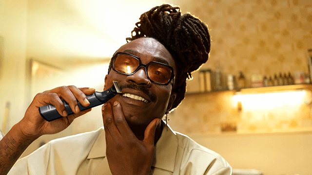 Een persoon met een snor en dreadlocks gebruikt een zwarte trimmer op zijn gezicht in een badkamer met een plank en flessen.