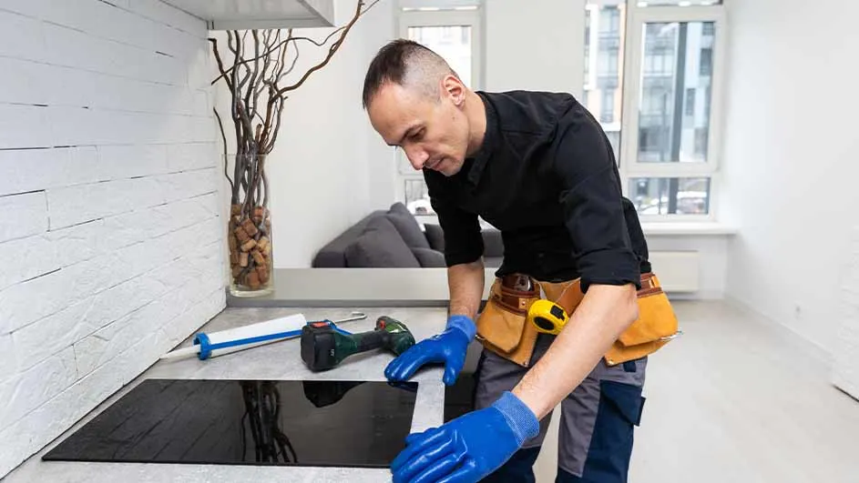 Een man met blauwe handschoenen installeert een zwarte kookplaat. Gereedschap, kit en een vaas staan op het aanrecht.