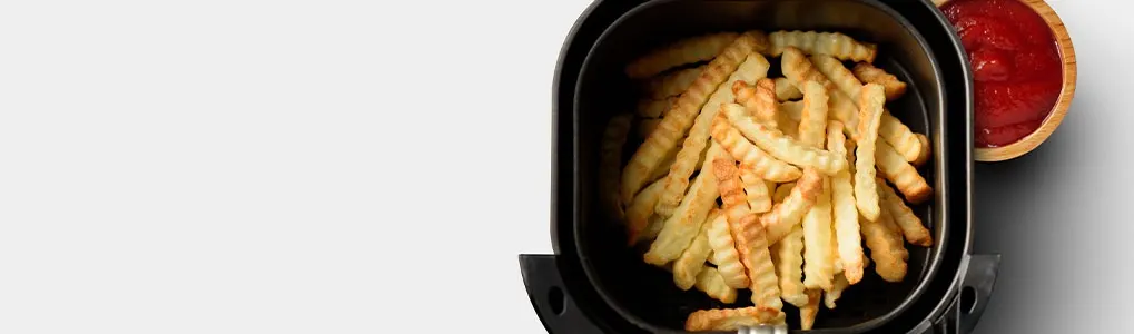 French fries fill a black air fryer basket, near a wooden bowl with red ketchup, on a white surface.