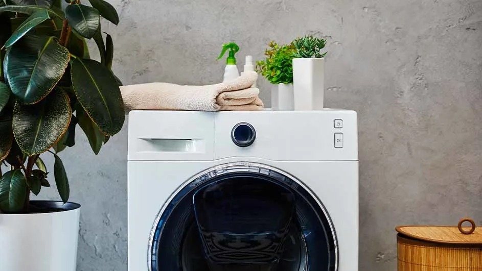 A white washing machine with a towel, spray bottle, and two potted plants is set against a gray wall.