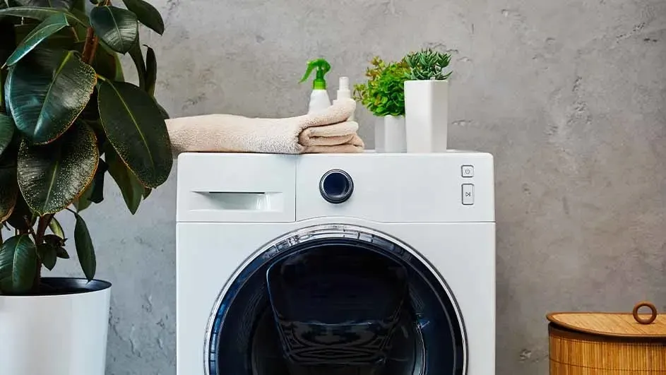A white washing machine with a towel, spray bottle, and two potted plants is set against a gray wall.