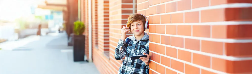 A person wearing headphones and a plaid shirt, holding a phone, stands by a brick wall on an outdoor pathway.