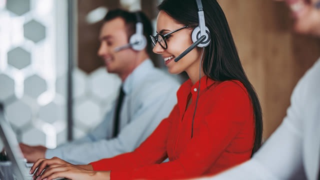 Customer service representatives wearing headsets work on computers in a call center, woman in red blouse in foreground.