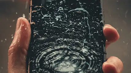 Een hand houdt een zwarte smartphone vast die bedekt is met waterdruppels.