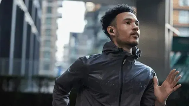 Man running through a city in a rain jacket with wireless earbuds, focused amid tall buildings.