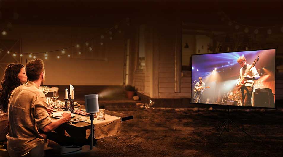A couple dine outdoors under warm lights while watching a concert on a large screen in the evening.