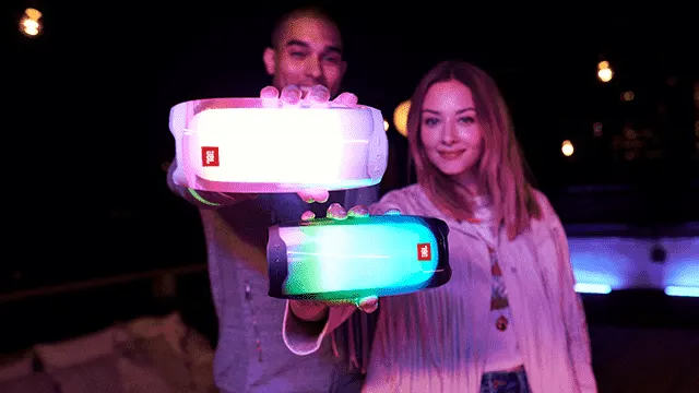 Twee mensen houden verlichte rechthoekige speakers vast met rode logo's tegen een donkere achtergrond.