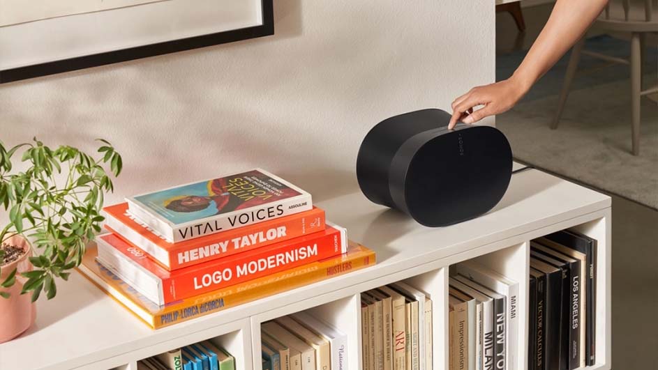 Boekenplank met zwarte speaker, hand bedient knop. Plant, ingelijste print, en boekenstapel met titels zoals 'Vital Voices'.