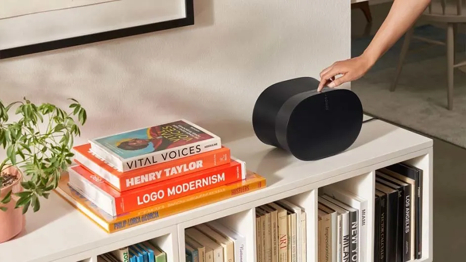 Boekenplank met zwarte speaker, hand bedient knop. Plant, ingelijste print, en boekenstapel met titels zoals 'Vital Voices'.