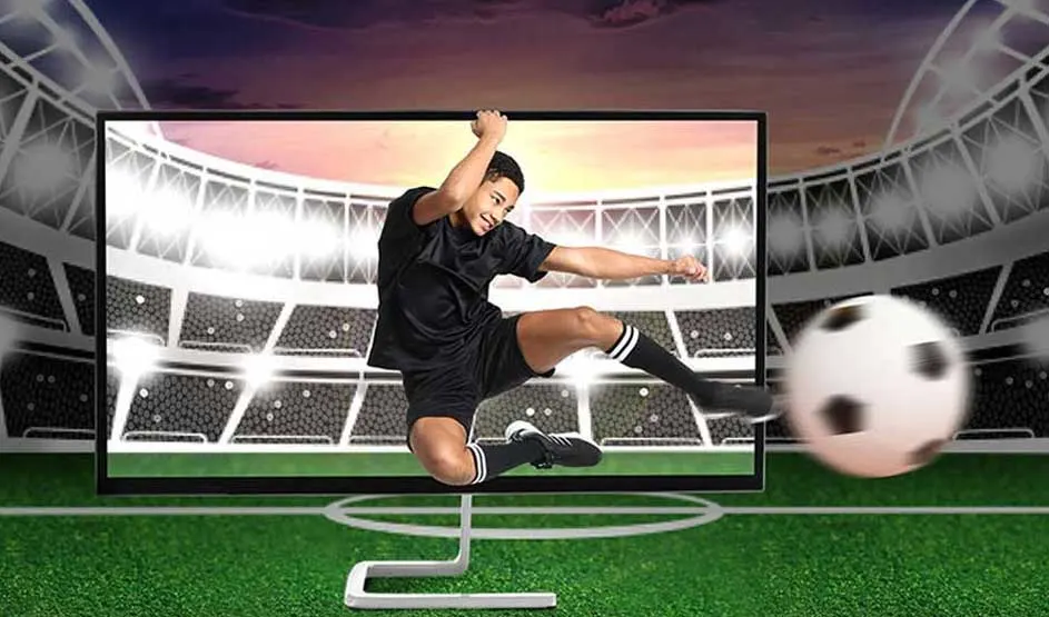 A footballer in black shoots at goal in a stadium; the ball flies large and blurred towards the viewer.