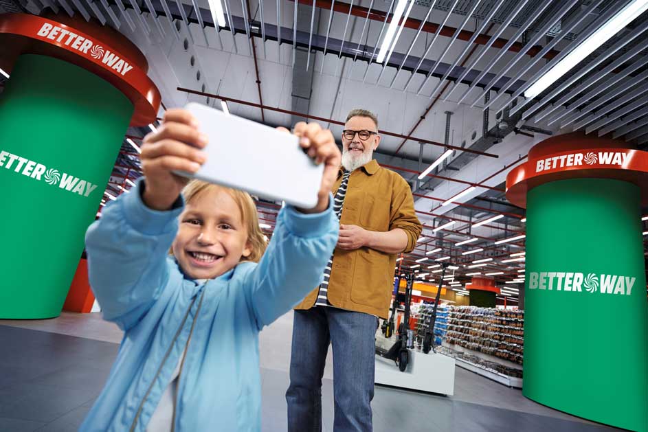 Een glimlachend kind maakt een selfie in een winkel, terwijl een oudere man erachter staat tussen grote BetterWay-zuilen.