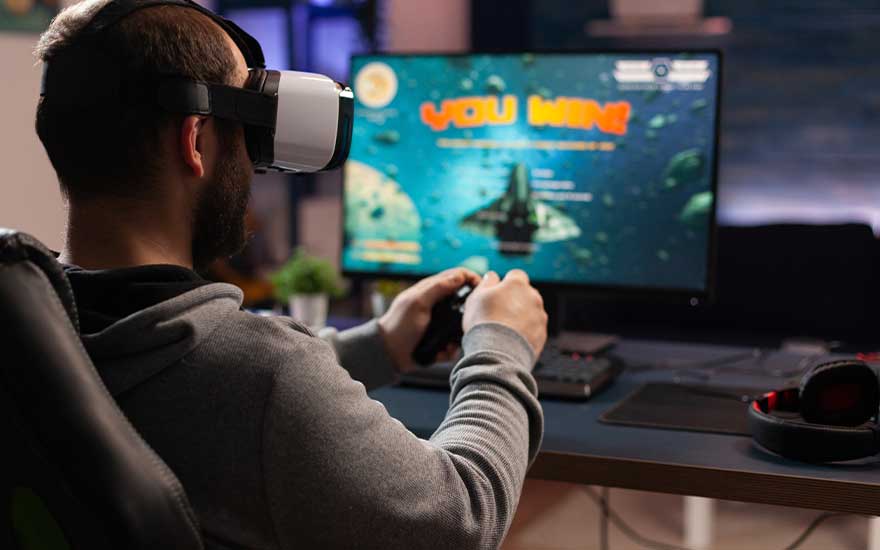 Een man speelt met een VR-bril en controller, zijn scherm toont "YOU WIN" terwijl hij een ruimtespel speelt aan zijn bureau.