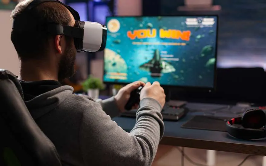 Een man speelt met een VR-bril en controller, zijn scherm toont "YOU WIN" terwijl hij een ruimtespel speelt aan zijn bureau.