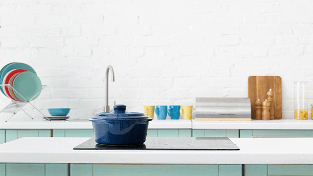 Een blauwe kookpan op een zwarte kookplaat, met een witte stenen muur en keukengerei op de achtergrond.