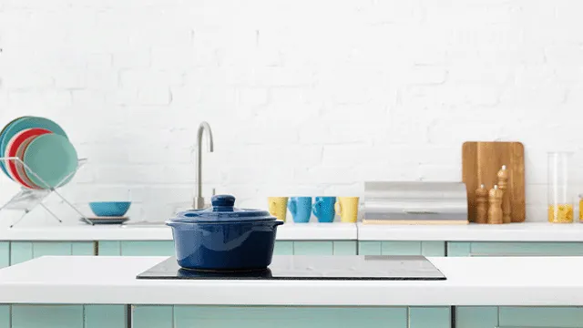 Een blauwe kookpan op een zwarte kookplaat, met een witte stenen muur en keukengerei op de achtergrond.