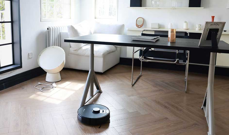 Een robotstofzuiger staat bij een bureau in een lichte woonkamer, met witte bank, radiator, raam en accessoires op een wandplank.