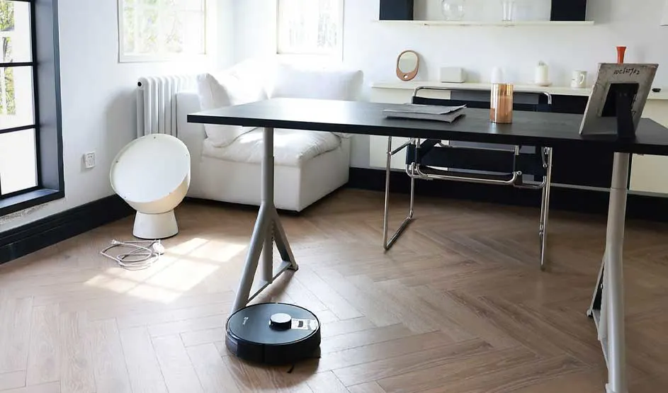 Een robotstofzuiger staat bij een bureau in een lichte woonkamer, met witte bank, radiator, raam en accessoires op een wandplank.