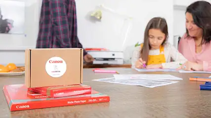 Een tafel met Canon-producten, een kind en een volwassene die aan het tekenen zijn, en een printer op de achtergrond.