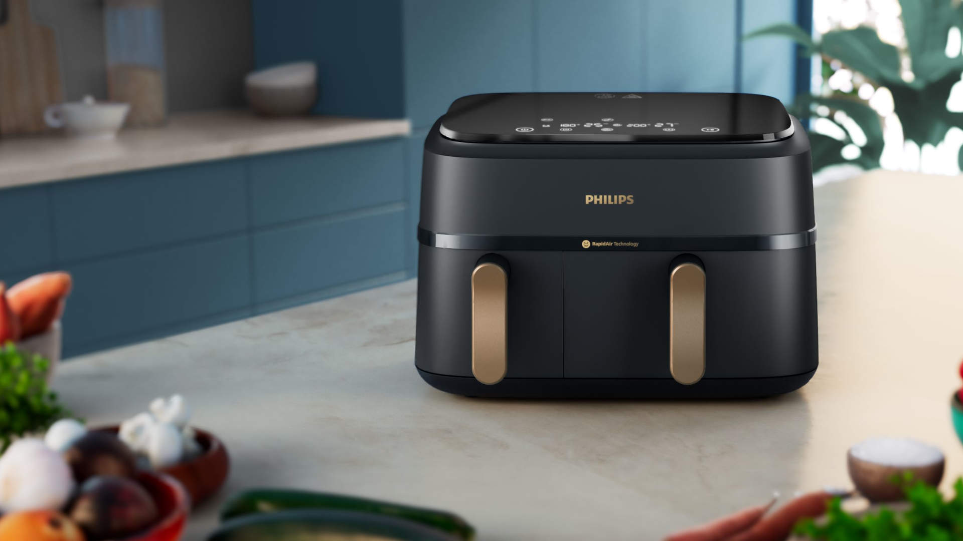 Een zwarte Philips airfryer staat op een aanrecht, met groenten op de voorgrond en blauwe kasten op de achtergrond.