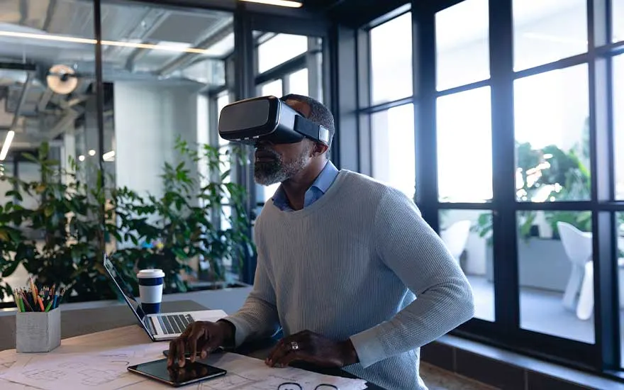 Een oudere man werkt aan een bureau met VR-headset, gebruikt een tablet terwijl hij omringd is door kantoorplanten en zonlicht.