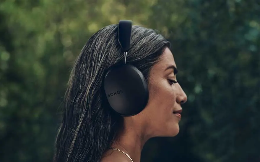 Woman with closed eyes enjoys music in a lush garden, wearing large black headphones and standing peacefully in profile view.