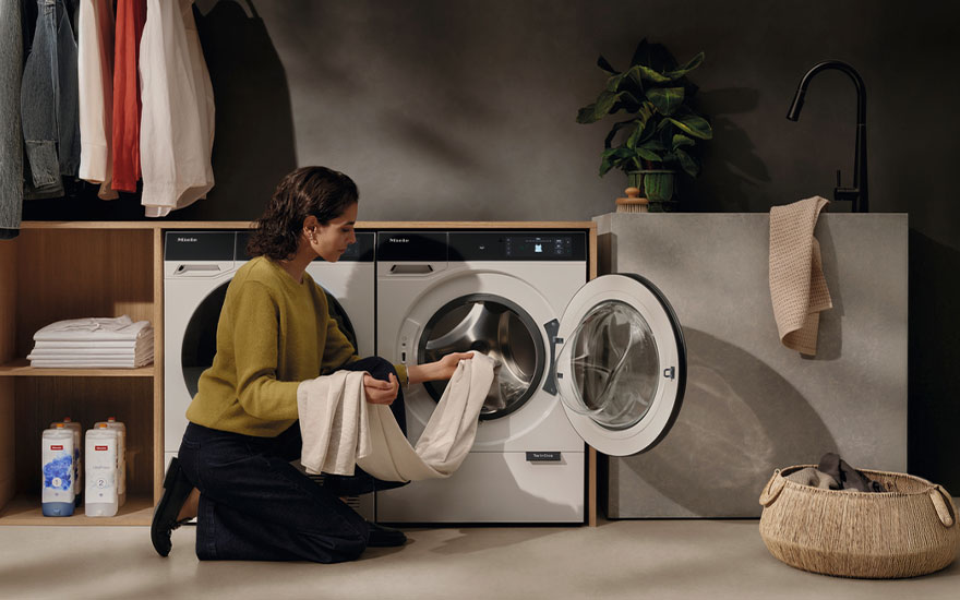 Moderne wasruimte met vrouw die witte was in Miele wasmachine doet, met houten opbergruimte en rieten wasmand. 