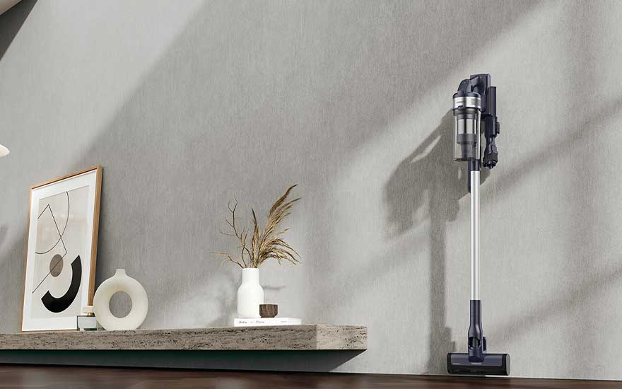 Een zilvergrijze steelstofzuiger hangt rechtop aan de muur naast een minimalistisch dressoir met kunstwerk, vazen en gedroogde grassen.