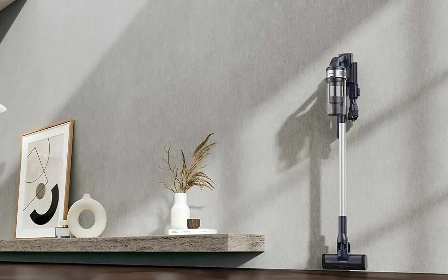 Een zilvergrijze steelstofzuiger hangt rechtop aan de muur naast een minimalistisch dressoir met kunstwerk, vazen en gedroogde grassen.