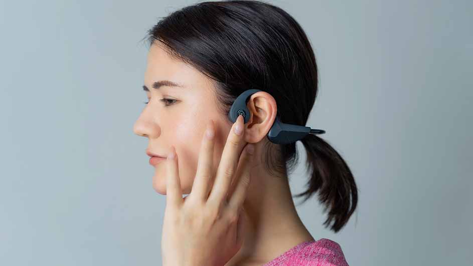 Een vrouw met een grijze hoofdtelefoon om haar oor, haar hand rustend op haar gezicht, tegen een grijze achtergrond.