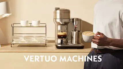 Koffiehoek met een Nespresso Vertuo-machine op een houten blad; een persoon houdt een cappuccino vast, met lamp en rek met kopjes op de achtergrond.