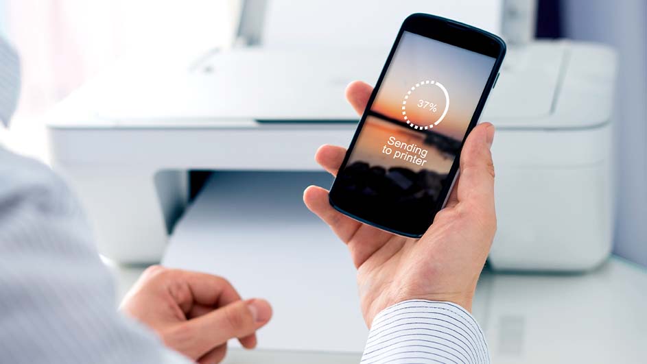 Hand houdt een telefoon vast met 'Sending to printer' tegen een witte printer op een witte tafel.