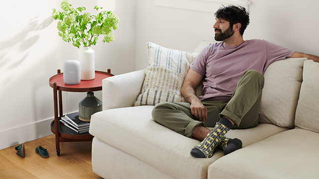 Een man zit op een witte bank in een kamer. Naast hem staat een rode tafel met een vaas bloemen en een speaker.