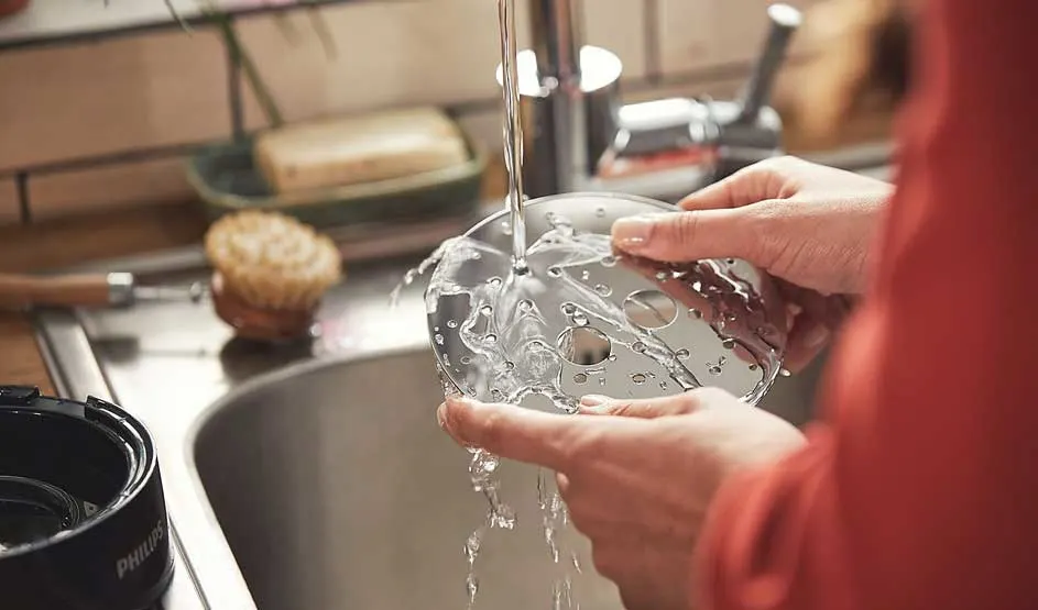 Iemand wast een metalen onderdeel van een Philips-apparaat onder de kraan in de keuken, met stromend water en een spons op de achtergrond.