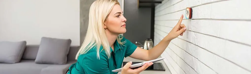 Vrouw bedient een slimme thermostaat aan de muur in een moderne keuken en houdt tegelijkertijd een tablet vast.