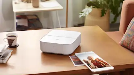 Witte printer op tafel met een telefoon, foto, koffiekop en documenten. Op de achtergrond een stoel en plant.