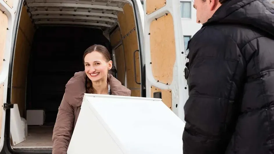 Een lachende vrouw en een man tillen samen een groot wit apparaat in of uit een bestelwagen, klaar voor verhuizen of levering.