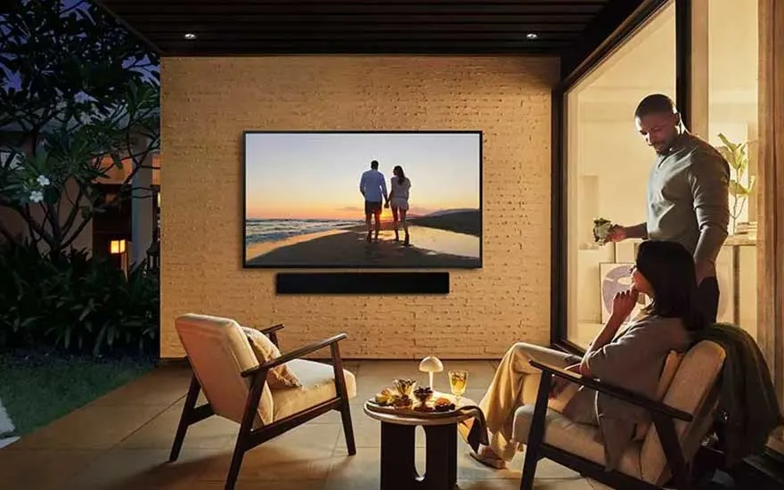 In a stylish patio setting, two people enjoy a relaxing moment while watching a large TV showing a sunset scene.