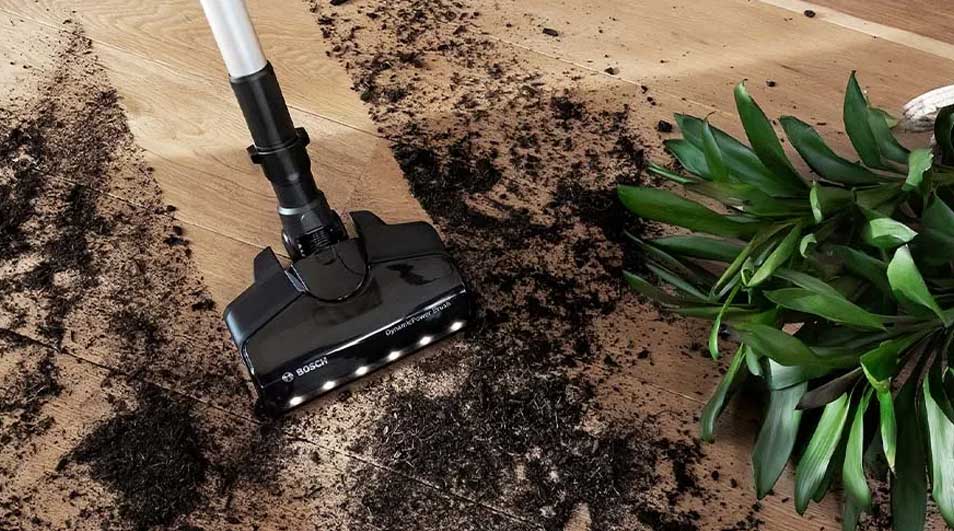 Een zwarte stofzuiger zuigt aarde op van een houten vloer naast een groene plant.
