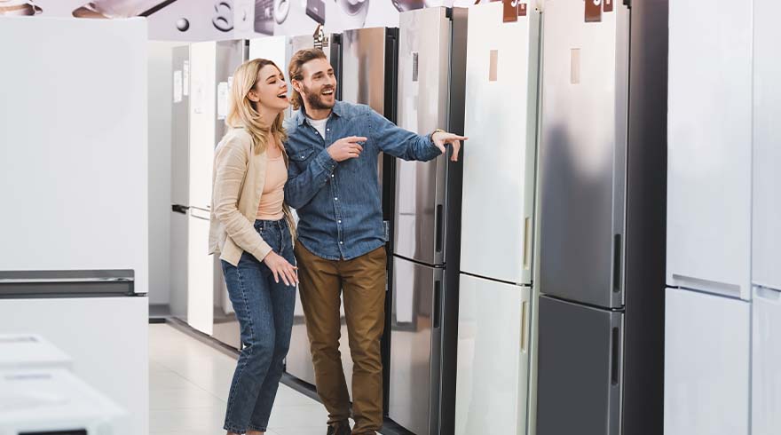 Een man en een vrouw bekijken koelkasten in een winkel; de man wijst naar een zilveren koelkast.