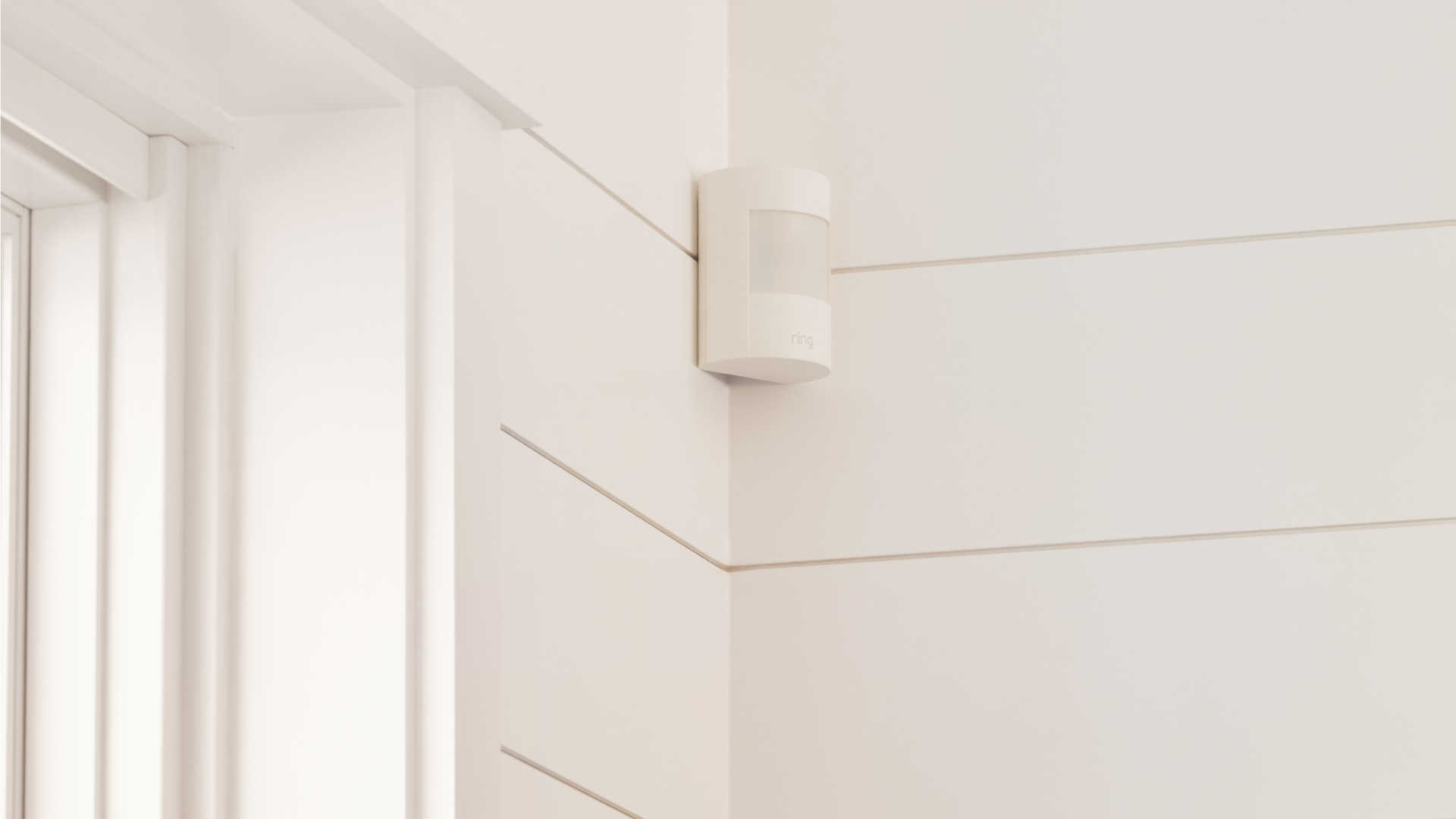 Witte bewegingssensor van Ring aan een witte lambrisering muur, nabij een witte deurpost.