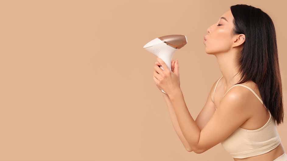 Een vrouw blaast naar een witte en koperen föhn, tegen een effen achtergrond. Ze draagt een topje met bandjes.