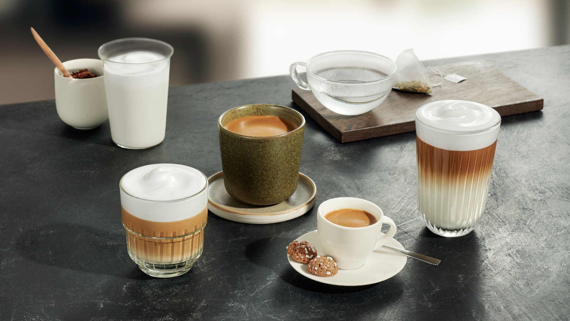 Diverse dranken staan op een donker oppervlak, waaronder koffie, melk en thee, in verschillende glazen en kopjes.