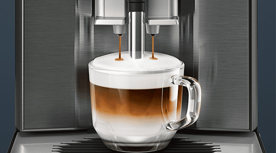 Koffiemachine vult een glazen mok met gelaagde koffie en melk. De machine is grijs en de achtergrond is donker.