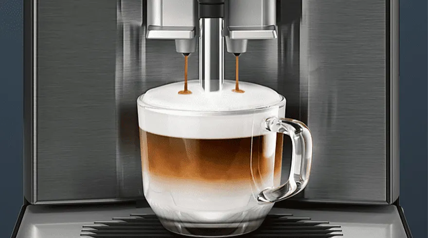 Koffiemachine vult een glazen mok met gelaagde koffie en melk. De machine is grijs en de achtergrond is donker.