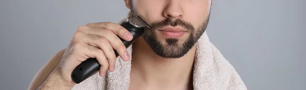 A person is using an electric razor to trim their beard, with a towel draped around their neck.