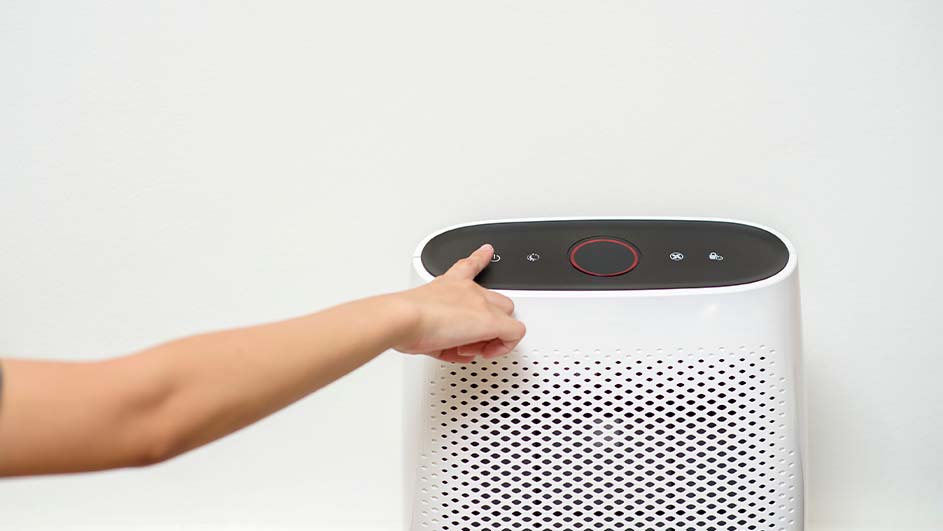 Een vinger drukt op de aan/uit-knop van een witte luchtreiniger met een zwart bedieningspaneel tegen een witte achtergrond.