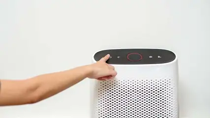 Een vinger drukt op de aan/uit-knop van een witte luchtreiniger met een zwart bedieningspaneel tegen een witte achtergrond.