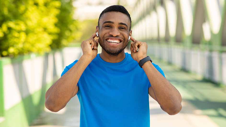 Een man lacht, draagt een blauw shirt, een horloge en witte oortjes. Achter hem een brug met groene bomen.